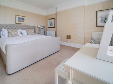 A bedroom with a bed, bedside table with lamp, framed pictures, and a white dressing table with chair at The Haven in Daymer Bay