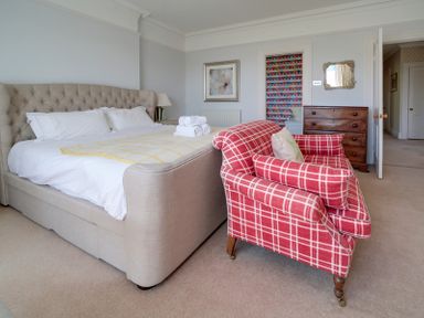 A bedroom with a beige upholstered bed white bedding a red plaid armchair and a wooden chest of drawers at The Haven in Daymer Bay
