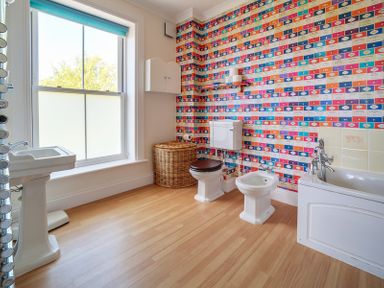 A bathroom with a large window a white sink a wicker laundry basket a toilet a bidet a bathtub and colorful wallpaper at The Haven in Daymer Bay