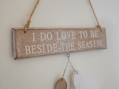 A wooden sign hanging on a wall with text I DO LOVE TO BE BESIDE THE SEASIDE and two decorative items hanging below at Pendaveli in Polzeath
