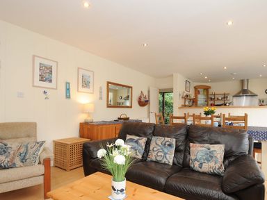 A living room with a black leather sofa and floral cushions a beige armchair a wooden coffee table with flowers and an open kitchen with a dining area at Pendaveli in Polzeath