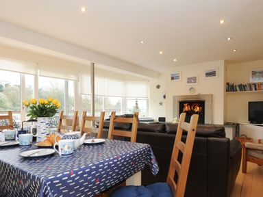 A dining table with cups and a vase of yellow flowers near windows and a living area with a fireplace at Pendaveli in Polzeath