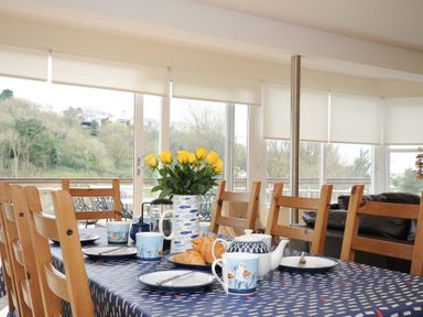 A dining table set with plates cups a teapot croissants and a vase with yellow roses in a room with wooden chairs and large windows at Pendaveli Polzeath