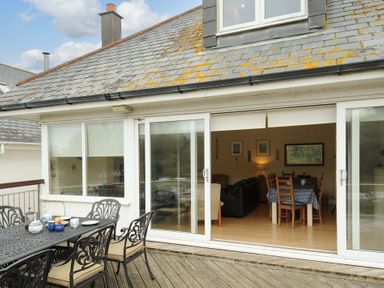 An outdoor dining area with a metal table and chairs on a wooden deck outside a house with sliding glass doors at Pendaveli in Polzeath