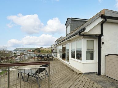 A large wooden deck with outdoor chairs and a dining table attached to a white house with large windows and a view of the sea at Pendaveli in Polzeath