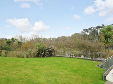 A lawn area with a wooden fence shrubs and railings overlooking trees at Pendaveli in Polzeath