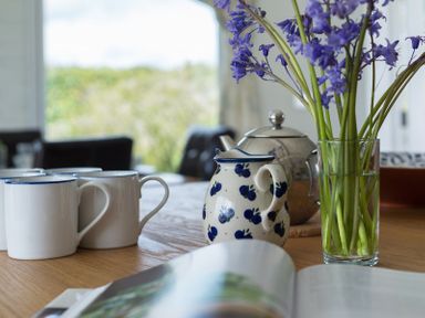 A wooden table with white mugs a patterned jug a metal teapot and a glass vase with purple flowers at Penny Liggan Rock