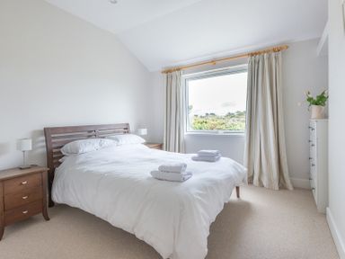 A bedroom with a double bed with white bedding two bedside tables with lamps a window with curtains and a chest of drawers with flowers at Penny Liggan in Rock