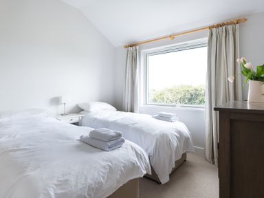 A bedroom with two single beds white bedding towels on beds a window and a wooden chest of drawers with flowers at Penny Liggan in Rock