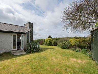 A garden with a lawn shrubs trees and a green shed next to a house with an open door at Penny Liggan in Rock