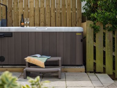 An outdoor hot tub with a small step bench holding a blanket and an open book with a wooden fence behind and greenery at Westray House in Polzeath