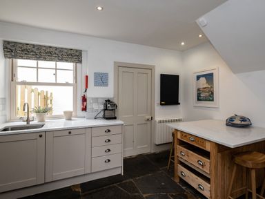 A kitchen with a countertop, sink, coffee machine, wooden island with drawers, stools, window, and wall art at Westray House in Polzeath