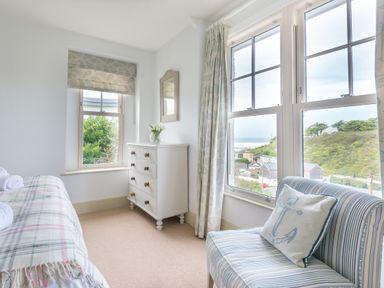 A bedroom with a striped chair a chest of drawers a window with a view and a bed with a blanket at Westray House in Polzeath