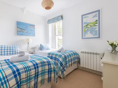 A bedroom with two single beds with blue checkered blankets and pillows a window a lamp and framed pictures at Westray House in Polzeath
