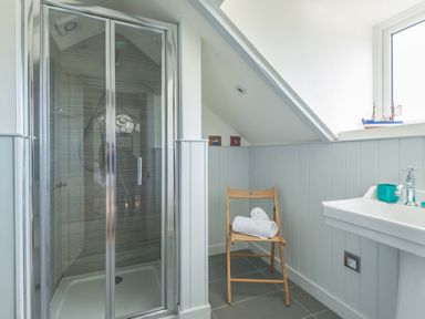 A bathroom with a glass shower enclosure a wooden folding chair with towels and a white sink under a window at Westray House in Polzeath