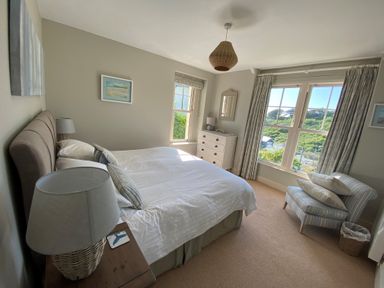 A bedroom with a double bed bedside tables lamps a chest of drawers an armchair and windows showing greenery outside at Westray House in Polzeath