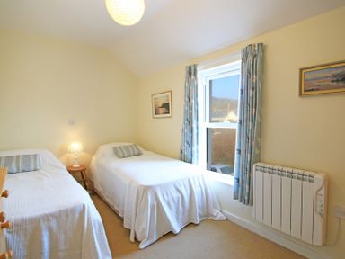 A bedroom with two single beds a bedside table with a lamp a window with curtains and two framed pictures on the walls at The Holiday House (2 Atlantic Mews) in Polzeath