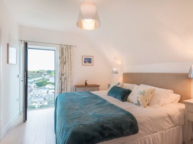 A bedroom with a bed with pillows and a throw with a door opening to a view of houses and hills at Stepper Point in Polzeath