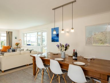A dining area with a wooden table, white chairs, plates, wine bottles, and flowers next to a living room with sofas and windows at Corriana in Rock