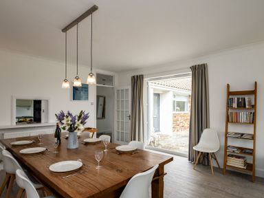 A dining room with a wooden table set with plates glasses and cutlery white chairs a vase with flowers a bookshelf and an open door leading outside at Corriana in Rock