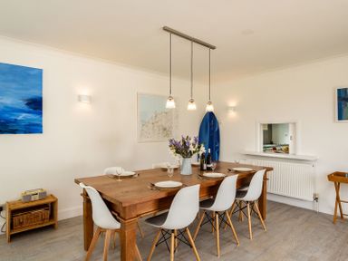 A dining room with a wooden table set for six white chairs hanging pendant lights and blue artwork on the walls at Corriana in Rock