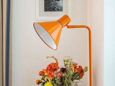 An orange floor lamp beside a bouquet of flowers with a framed picture on a white wall at Corriana in Rock