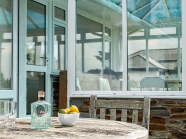 A wooden table with a glass bottle bowl of fruits and two glasses outside a glass conservatory at Corriana in Rock