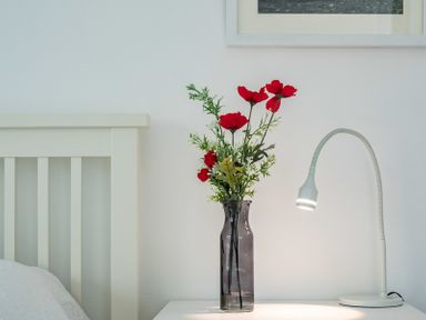 A bedside table with a vase of red flowers and a white reading lamp next to a bed at Corriana in Rock