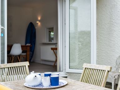 A wooden outdoor table with a tea set and chairs outside a house with open doors showing a dining area and a surfboard inside at Corriana in Rock