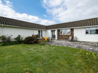 A single-story house with a tiled roof a stone patio area with chairs and a table and a grass lawn at Corriana Rock