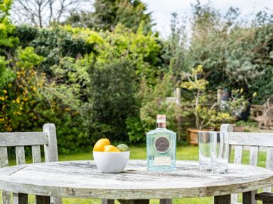 A round wooden table with a bowl of fruit a bottle of gin and two glasses in a garden with wooden chairs at Corriana in Rock