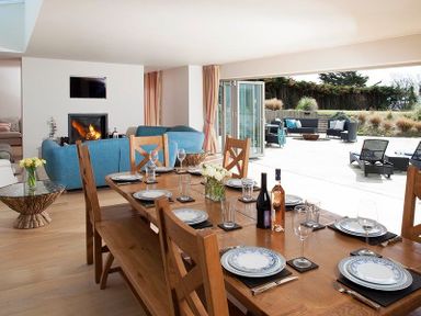 A dining area with a wooden table set with plates glasses and bottles next to a living room with blue sofas a fireplace and an open patio with outdoor seating at Downalong in Daymer Bay