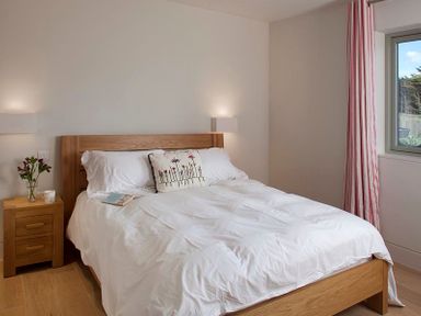 A bedroom with a wooden bed frame white bedding a floral cushion a wooden bedside table with flowers and a window with striped curtains at Downalong Daymer Bay