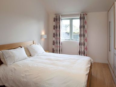 A bedroom with a wooden bed frame white bedding two pillows a window with patterned curtains and a wardrobe at Downalong in Daymer Bay