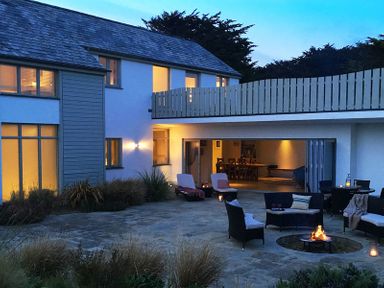 An outdoor patio with chairs around a fire pit and a dining area inside a modern house at Downalong in Daymer Bay