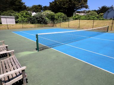 An outdoor tennis court with a net and wooden benches surrounded by trees and houses at Downalong Daymer Bay