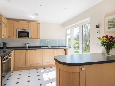 A kitchen with wooden cabinets a black countertop a stove a microwave a kettle flowers on a counter and glass doors overlooking a garden at Chy-An-Dowr in Rock