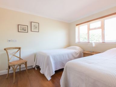 A bedroom with two single beds covered with white bedspreads a wooden chair two framed pictures on the wall and a window with blinds at Chy-An-Dowr in Rock