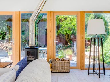 A living room with a wood stove and large windows showing a garden with chairs and plants at Greystones Studio in Daymer Bay