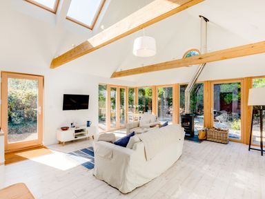 A living room with sofas a wood stove and large windows showing outdoor greenery at Greystones Studio in Daymer Bay