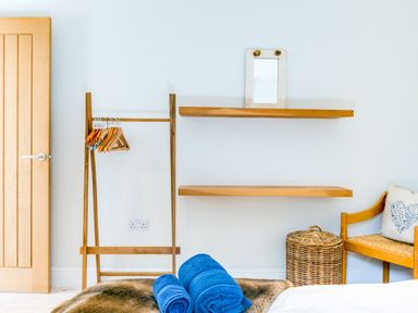 A bedroom corner with a wooden door wooden shelves wooden coat rack blue towels wicker basket and a wooden chair with a cushion at Greystones Studio in Daymer Bay