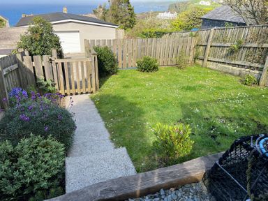 A small fenced garden with a grass lawn and a paved path leading to a wooden gate at The Porthole in Port Isaac