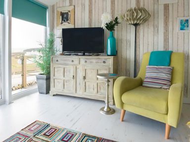 A living room with a green armchair and a striped pillow next to a wooden cabinet with a TV and a blue vase at The View in Port Isaac