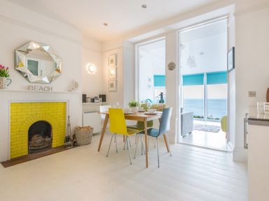A dining area with a wooden table and colorful chairs next to a yellow-bricked fireplace and windows showing an ocean view at The View in Port Isaac