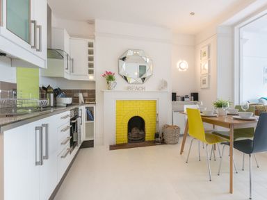 A kitchen with white cabinets a yellow tiled fireplace and a dining table with multicolored chairs at The View in Port Isaac