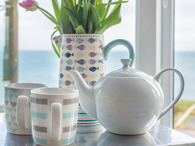 A teapot with two mugs and a pitcher with flowers on a table by a window with a seaside view at The View in Port Isaac