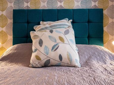 A bed with patterned cushions and a textured bedspread in front of a padded headboard and leaf patterned wallpaper at The View in Port Isaac