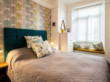 A bedroom with a bed featuring a teal headboard and patterned pillows beside a window with blinds and a built in seat at The View in Port Isaac