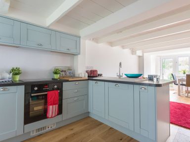 A kitchen with light blue cabinets black countertop and wooden floor adjacent to a dining area with a red rug at Beehive Cottage in Port Isaac