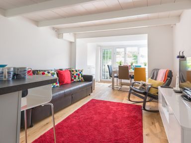 A living room with a brown sofa with colorful cushions a red rug and a dining table with colorful chairs at Beehive Cottage in Port Isaac